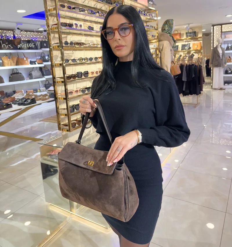 A guest holding a brown suede handbag in the store, with the sunglasses wall behind