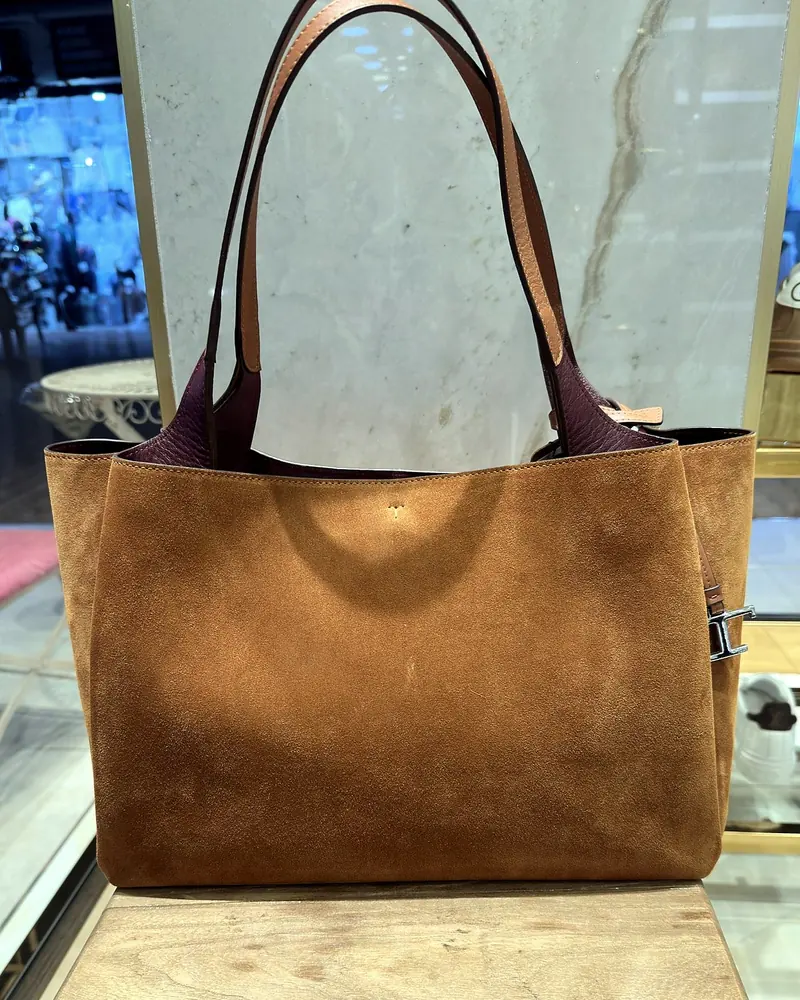 A tan suede leather tote with a burgundy lining, on the wooden stool in the BROTHERSBAG store