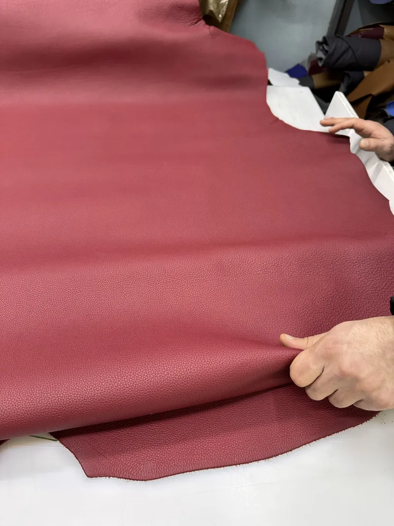 A hand bending a full burgundy hide in the workshop: real leather creases softly and recovers